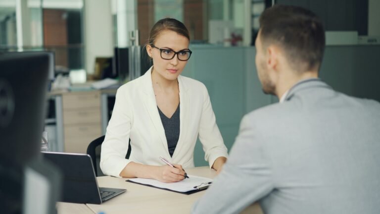 A woman in an office listens carefully while speaking with a colleague across a desk