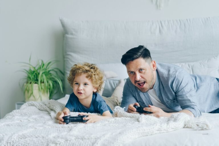 A father and young child playing video games together on a bed