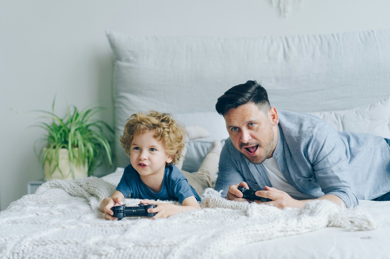 A father and young child playing video games together on a bed