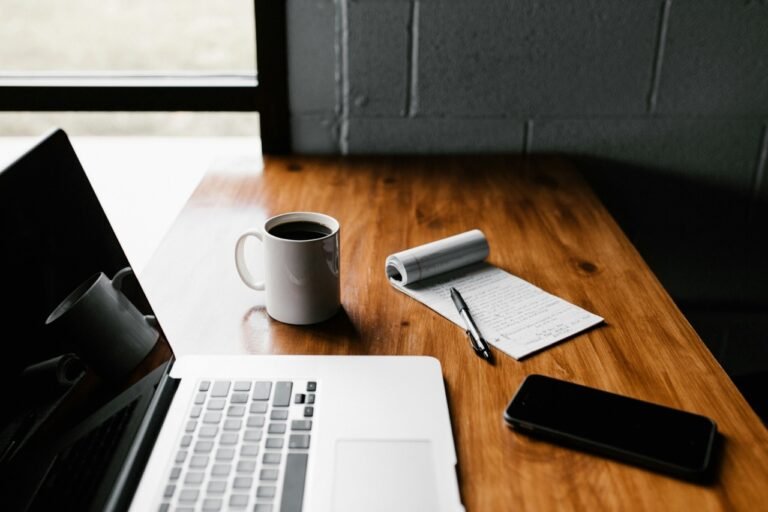 A clean workspace with a laptop, coffee mug, notebook, and smartphone on a wooden desk, representing modern work and productivity.