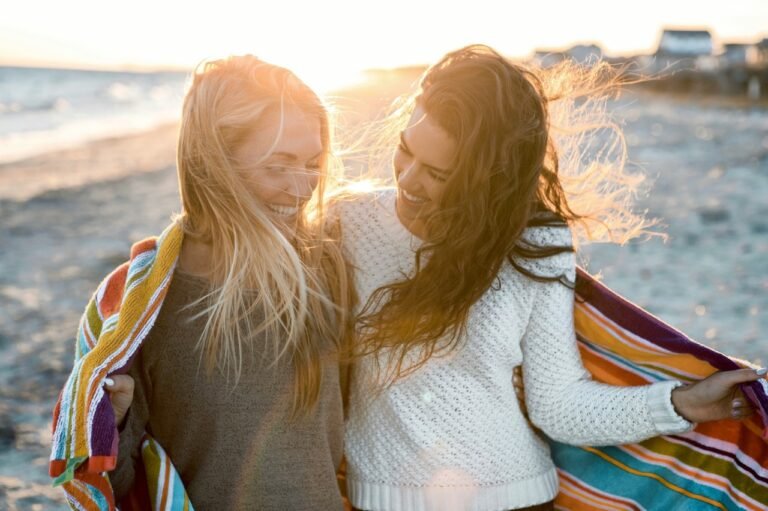 Two close friends laughing together at sunset on the beach, sharing a blanket