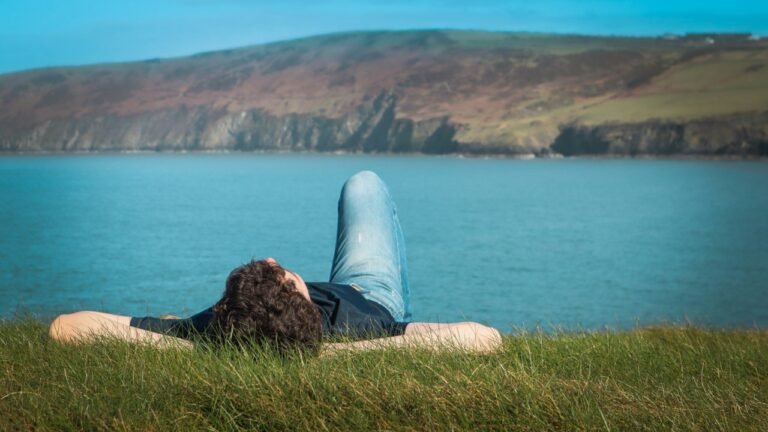 Man lying in the grass on a coastal hillside, resting and doing nothing while looking out at the sea