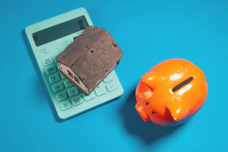 Small house model on a calculator beside a piggy bank, representing the financial decision of renting versus buying a home