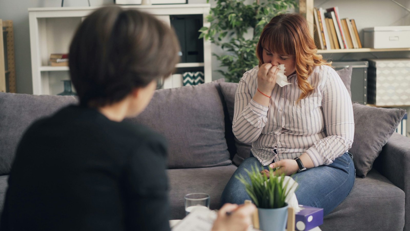 Woman blowing her nose into a tissue while sitting on a couch talking with someone