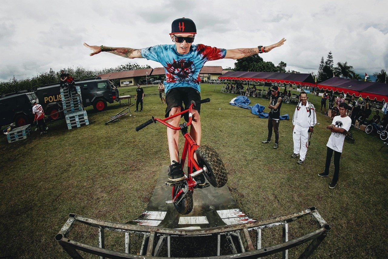 BMX rider performing a jump on a red bicycle at an outdoor event, with spectators watching in the background.