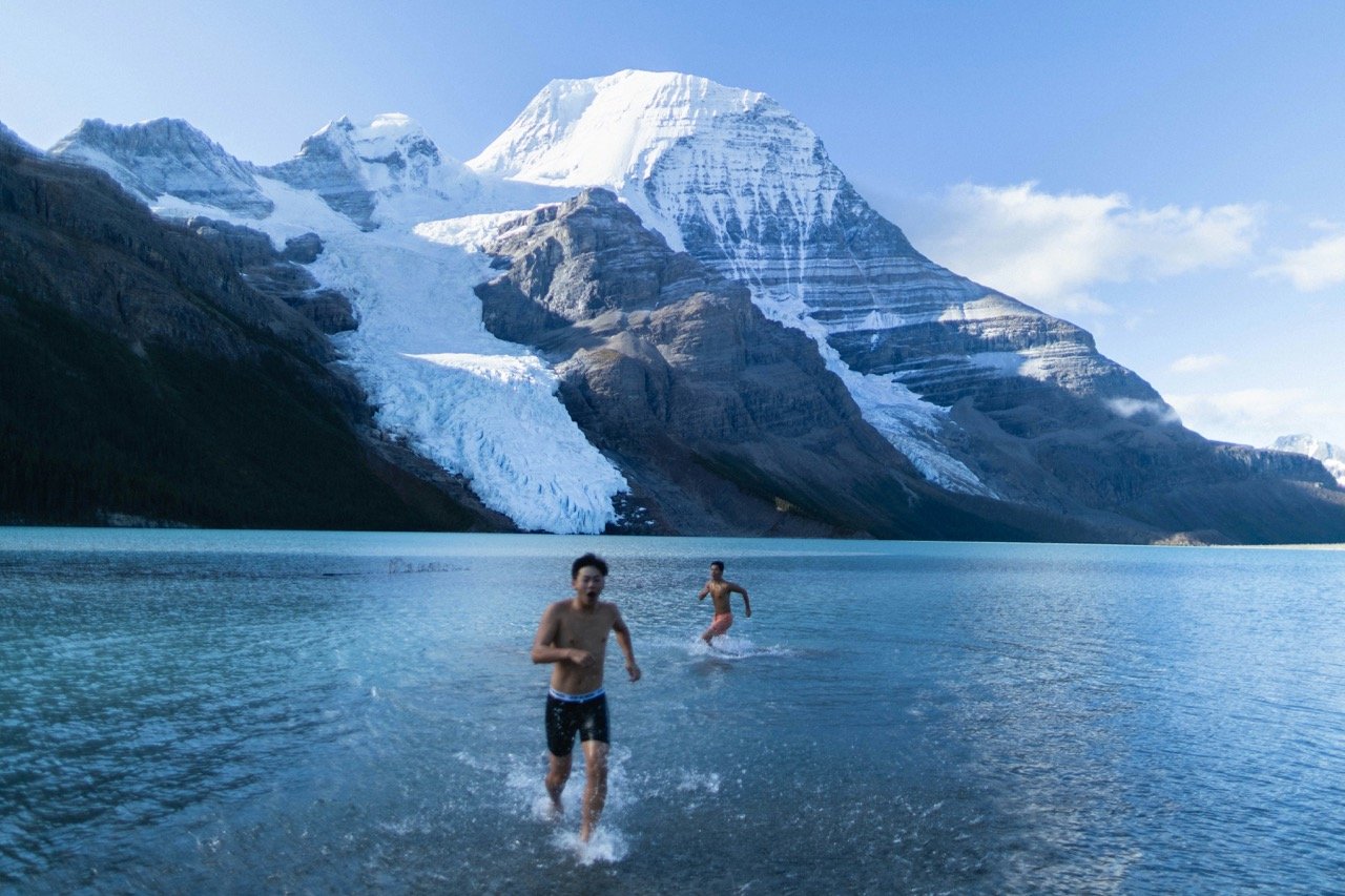 Two people swim in a cold mountain lake beneath snow-covered peaks and glaciers.