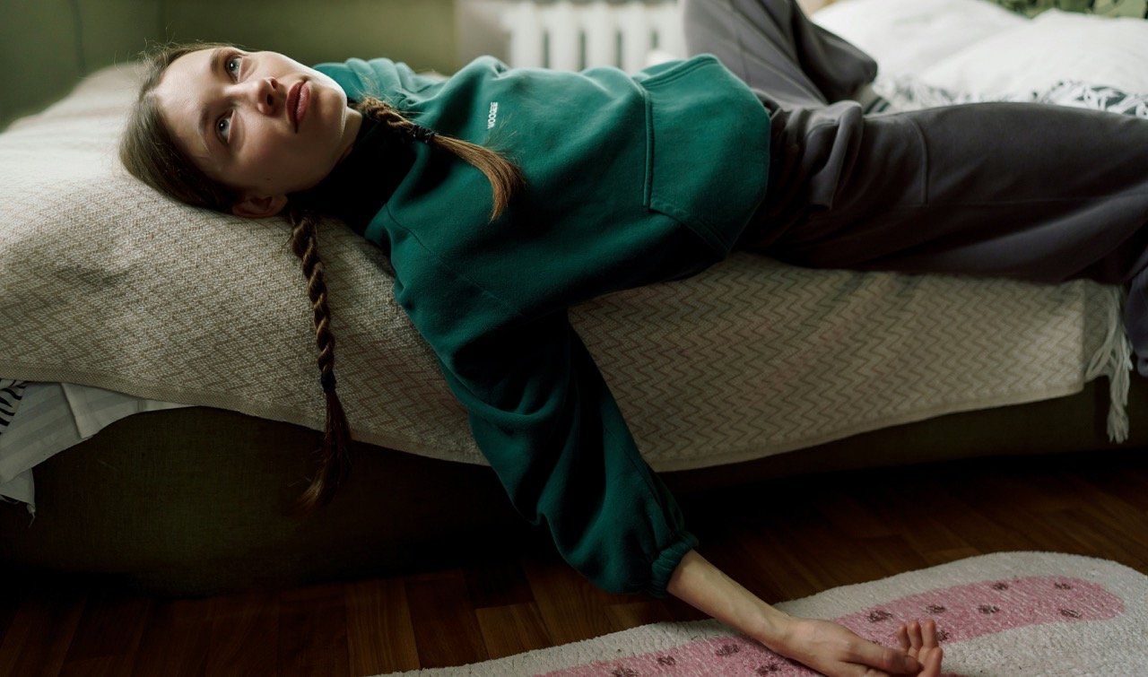 Woman lying across a bed with one arm reaching toward the floor, illustrating a quiet moment of rest and boredom.