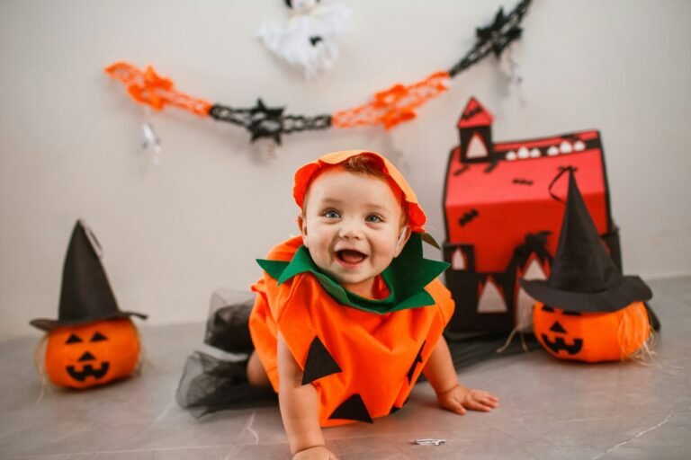 Smiling baby dressed in an orange pumpkin costume surrounded by Halloween decorations and jack-o'-lanterns.
