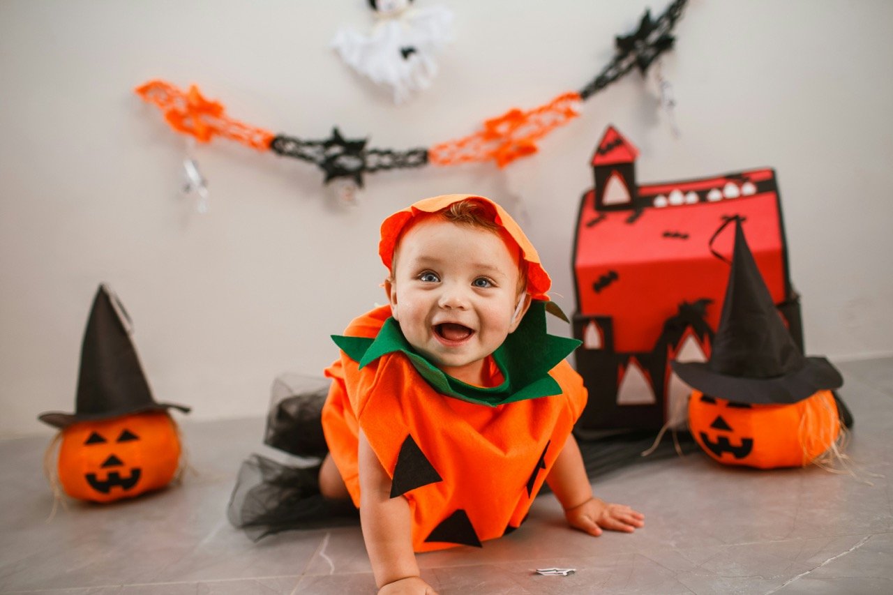 Smiling baby dressed in an orange pumpkin costume surrounded by Halloween decorations and jack-o'-lanterns.