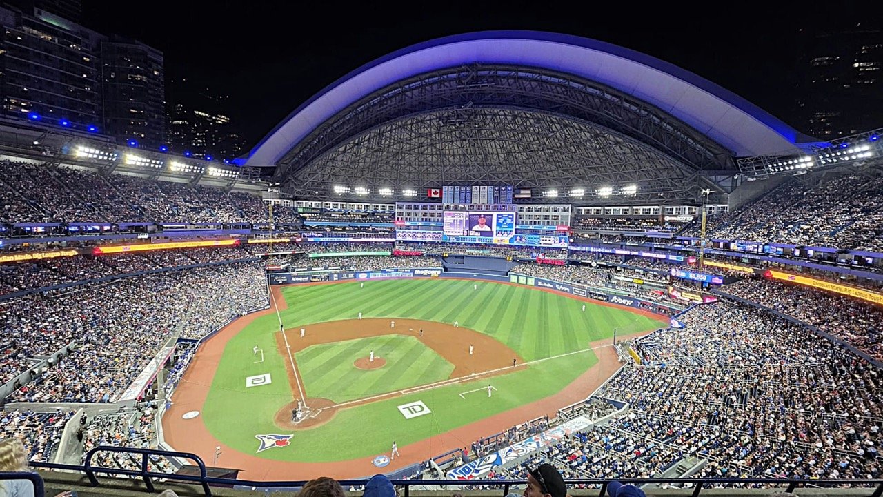 Packed baseball stadium during a nighttime game, with fans filling the stands around the brightly lit field.