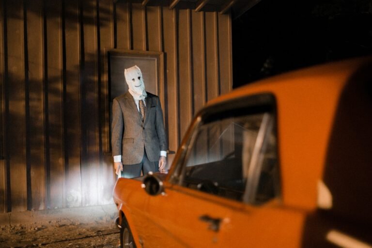 Person wearing a white mask and formal suit standing beside an orange car in a dimly lit, atmospheric setting.