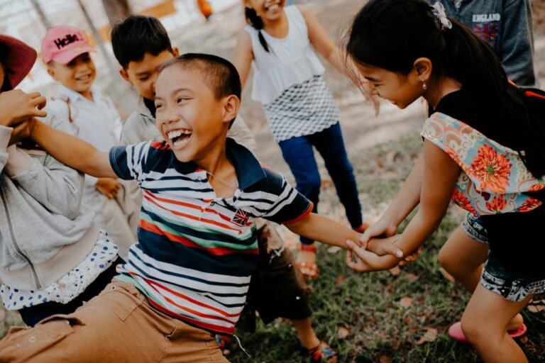 A group of children laughing and playing together outdoors, holding hands and enjoying a fun group activity.