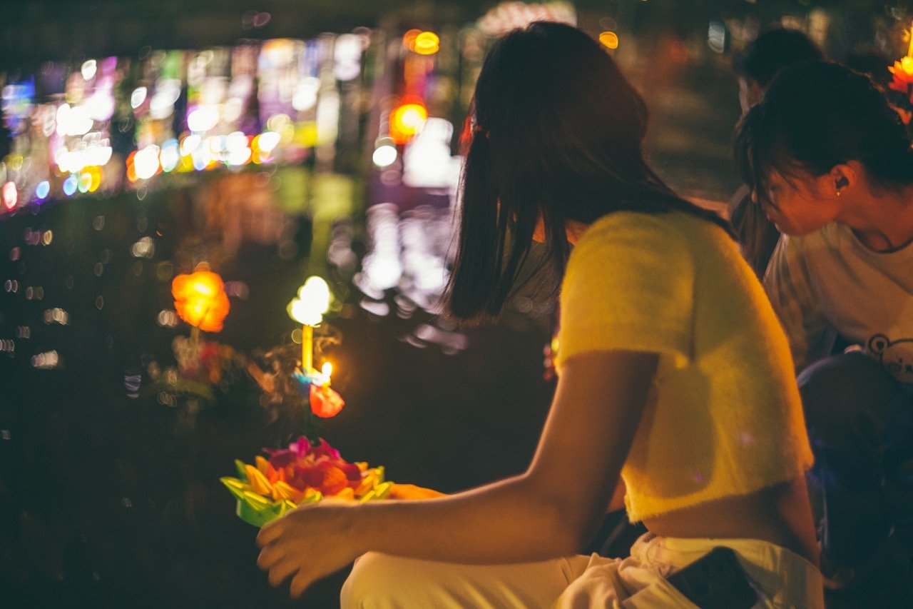 Woman preparing a floating krathong with flowers and candles on water at night