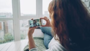 Woman sitting by a window and holding a smartphone during a video call with two smiling friends, staying in touch through long-distance friendship
